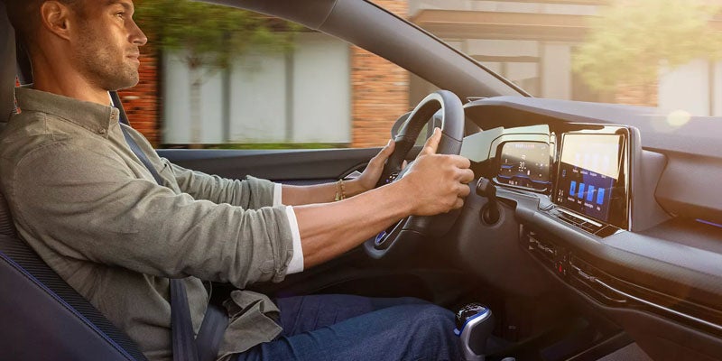 A man with short brown hair in a grey shirt and blue jeans driving a modern car, seen from the right.