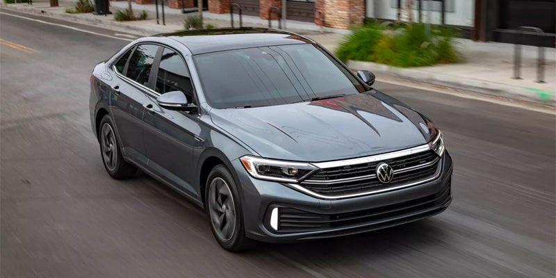 A dark gray Volkswagen Jetta sedan with a black roof drives on a street, angled toward the viewer.
