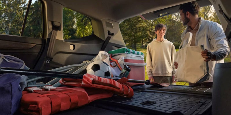 A man and a boy load various sports and camping gear into the back of a car with an open tailgate.