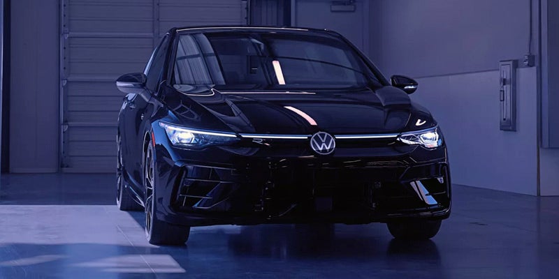 Front view of a sleek black hatchback car in a dark garage, headlights and light bar illuminated.