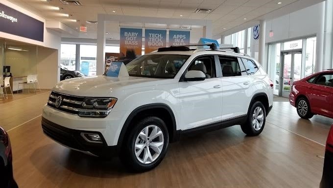 White Volkswagen Atlas SUV on display in a car dealership showroom with a red car and office in the background.
