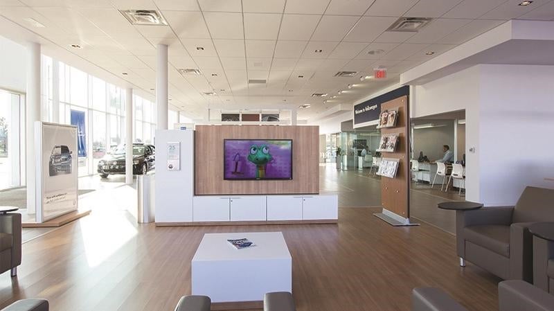 Modern car dealership waiting area with a TV showing a cartoon, seating, glass walls, and vehicles in the showroom.